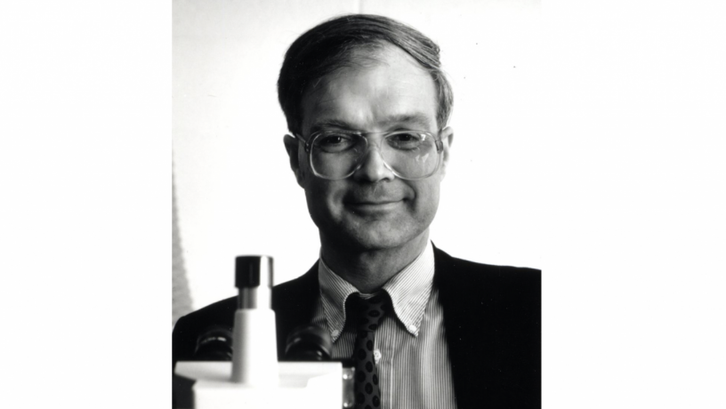 A middle aged white man is standing behind a microscope. He is wearing a suit coat and tie. 