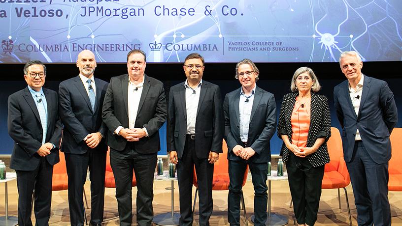Seven panelists, six male and one female, stand on stage to pose for a photo.