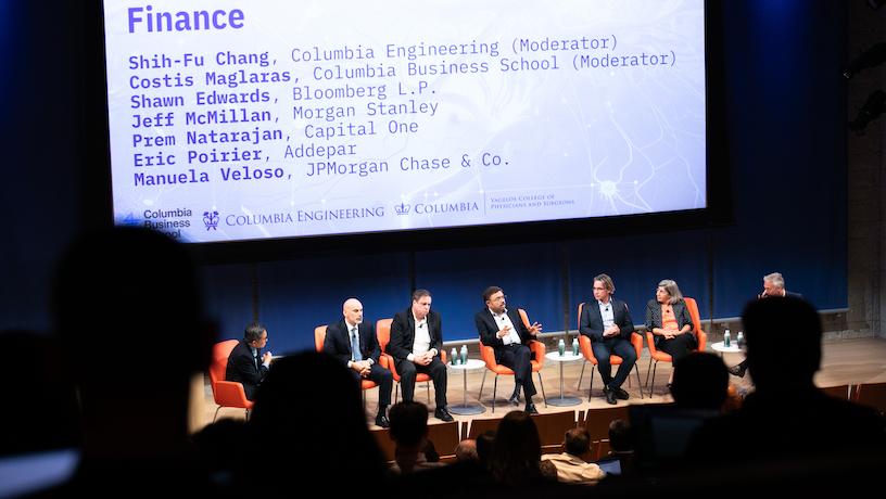 The audience watches the speaker panel as they sit on stage. The screen behind them reads their names: Shih-Fu Chang, Columbia Engineering (Moderator); Costis Maglaras, Columbia Business School (Moderator); Shawn Edwards, Bloomberg L.P.; Jeff McMillan, Morgan Stanley; Prem Natarajan, Captial One; Eric Poirier, Addepar; Manuela Veloso, JPMorgan Chase & Co.