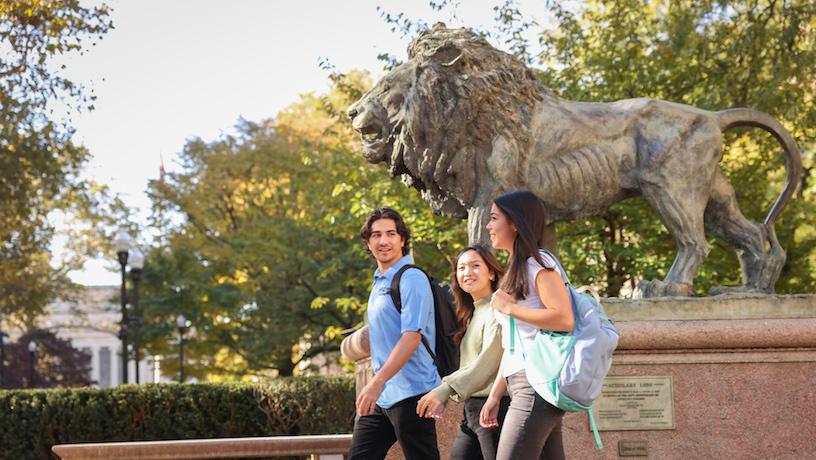 Students walking on Columbia University's tree-lined campus. A lion sculpture behind the students has plaque that reads "Scholar's Lion".