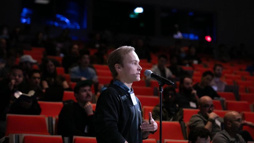 An attendee speaking into a microphone. Behind them audience members sit scattered in the auditorium.