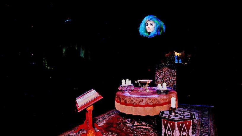 Madame Leota's floating head appears above a candle adorned table and a lectern with an open book.