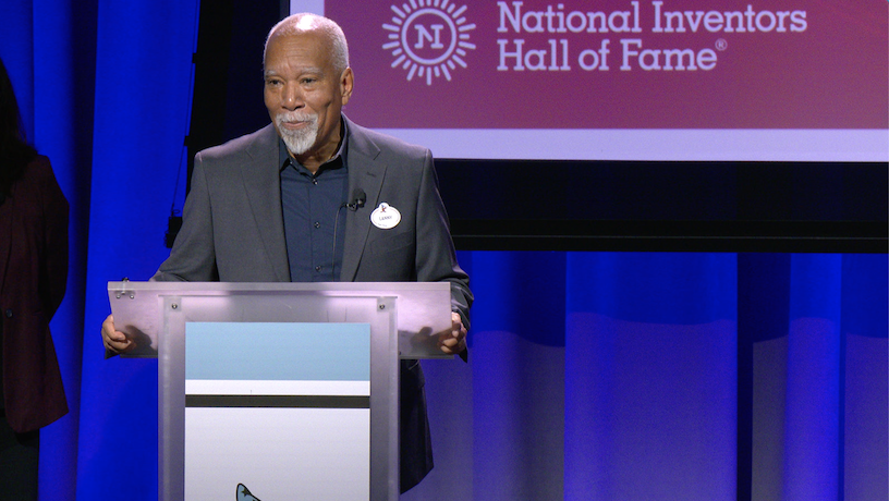 Lanny Smoot standing at a lectern. The screen behind him reads "National Inventors Hall of Fame".