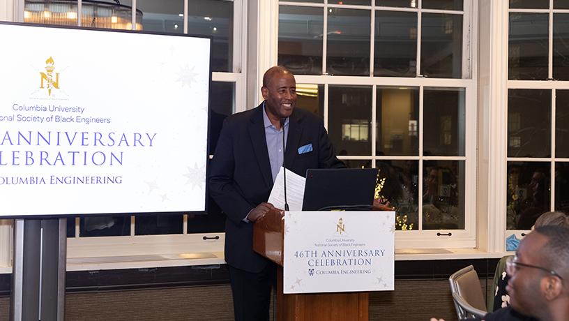 A smiling person standing at podium with a microphone. The podium sign and screen behind them reads "Columbia University | National Society of Black Engineers | 46th Anniversary Celebration"