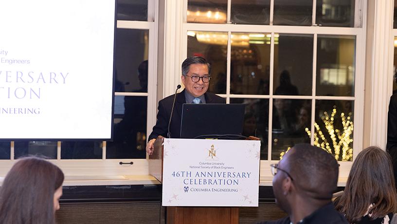 Columbia Engineering Dean standing at podium with a microphone. The podium sign and screen behind them reads "Columbia University | National Society of Black Engineers | 46th Anniversary Celebration"