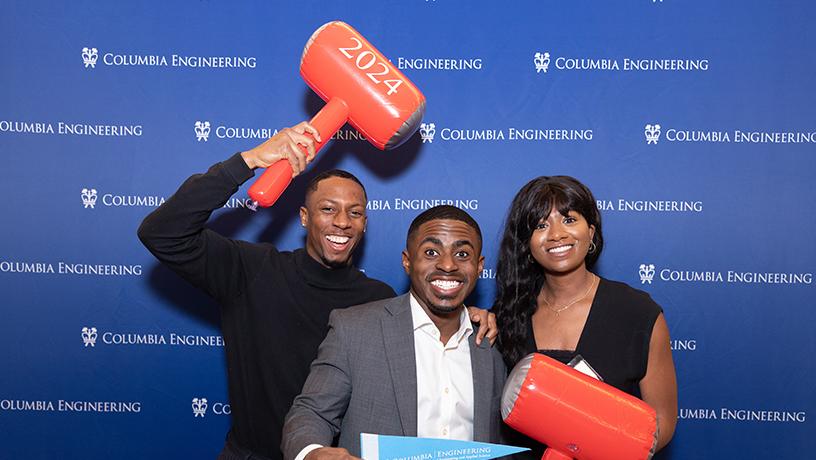 Three people posing for a photo. The left and right-most people hold inflatable hammers. The middle person holds a Columbia Engineering pennant. The photo background shows the Columbia Engineering logo.