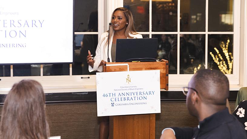 A smiling person standing at podium with a microphone. The podium sign reads "Columbia University | National Society of Black Engineers | 46th Anniversary Celebration"