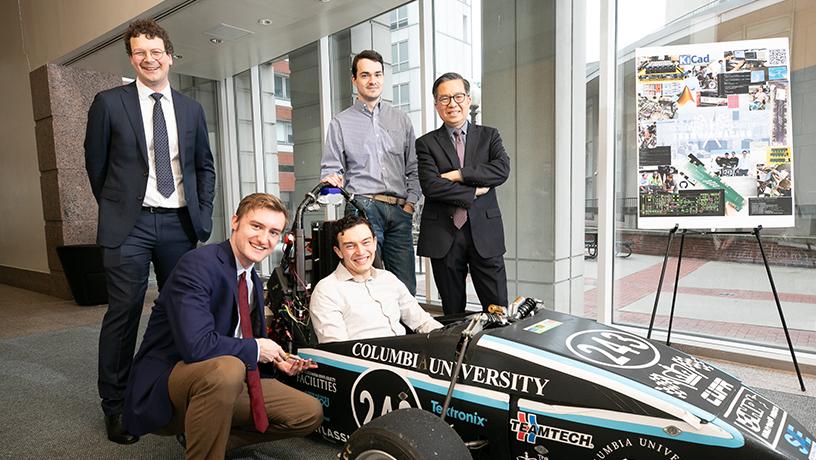 Four people standing around A Columbia Formula Racing student club's electric vehicle. One person sits in the vehicle's drivers seat.