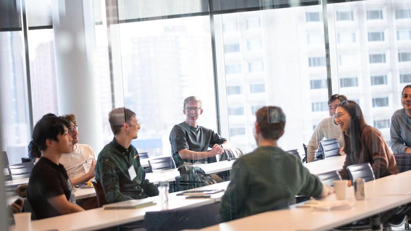 Eight attendees sit at desks conversing with eachother.