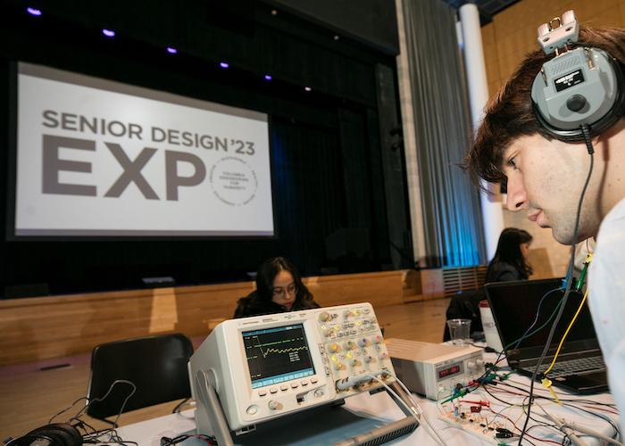 A student wearing headphones, a sign reads "Senior Design '23 Expo"