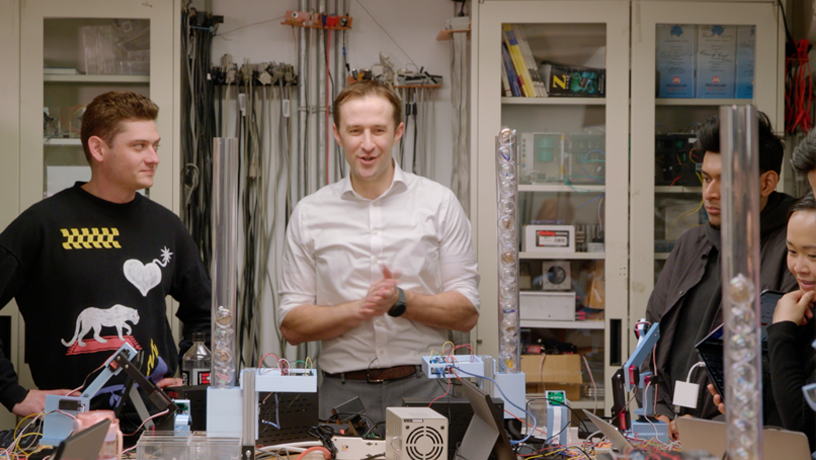 A professor with their hands together standing in a room cluttered with wires and electronics. Students look at them from either side.