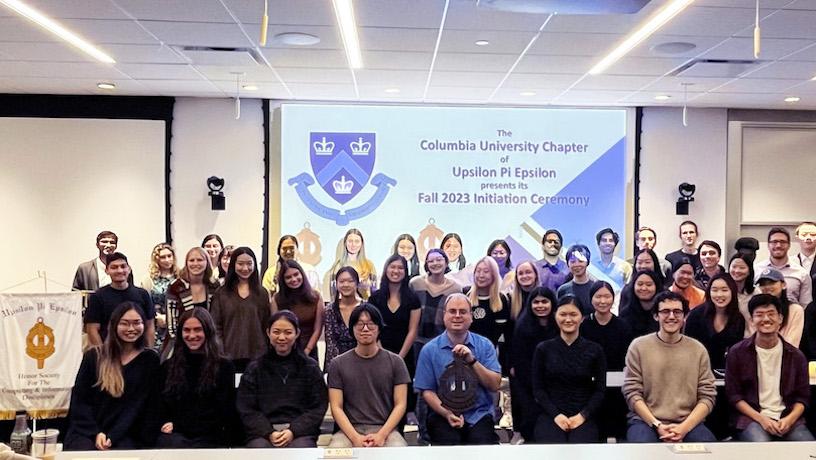 Three rows of people smiling for a photo. In the background, a large screen reads "Columbia University Chapter of Upsilon Pi Epsilon Fall 2023 Initiation Ceremony