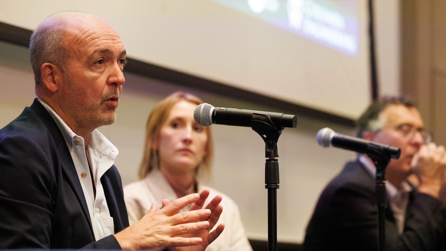 A panelist speaking into a microphone. Two other panelists appear out of focus looking at the speaker