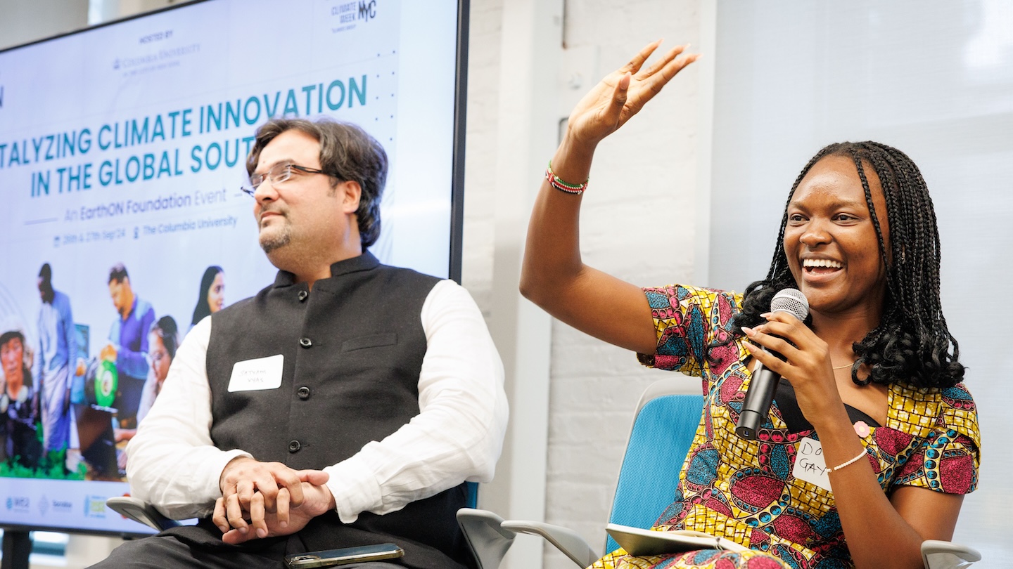 Two people sitting. The one on the right holds a microphone while raising their hand over their head.
