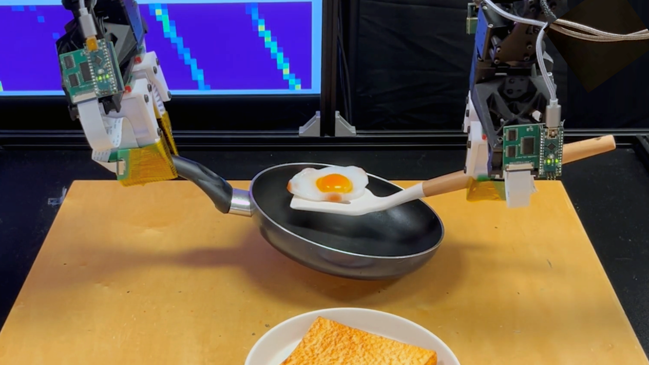 Two robotic hands. The one the left holds a pan. The one on the right balances a fried egg with a spatula.