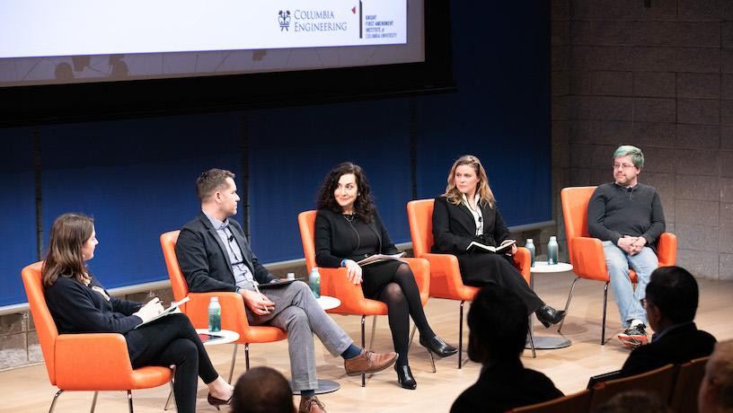 Five panelists sitting in a row on-stage. The backs of audience members' heads appear in the foreground.