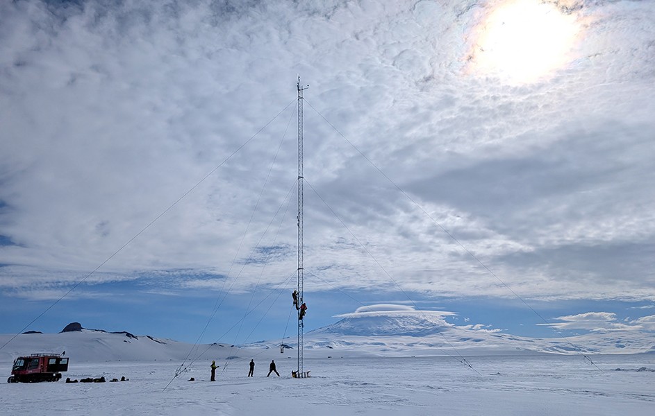 Two people climb a radio tower in a snowy, antarctic landscape