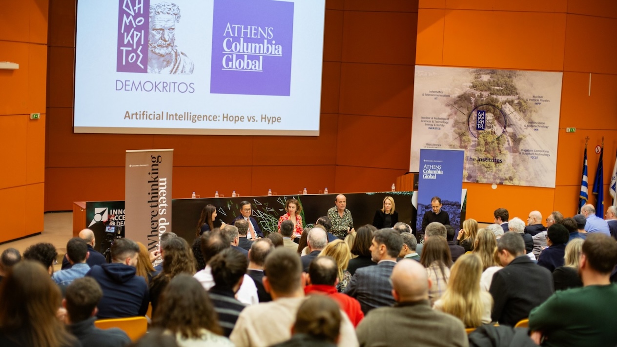 A panel of six people in a crowded auditorium. The projector screen above the panel reads "Athens Columbia Global; Artificial Intelligence: Hope vs. Hype""