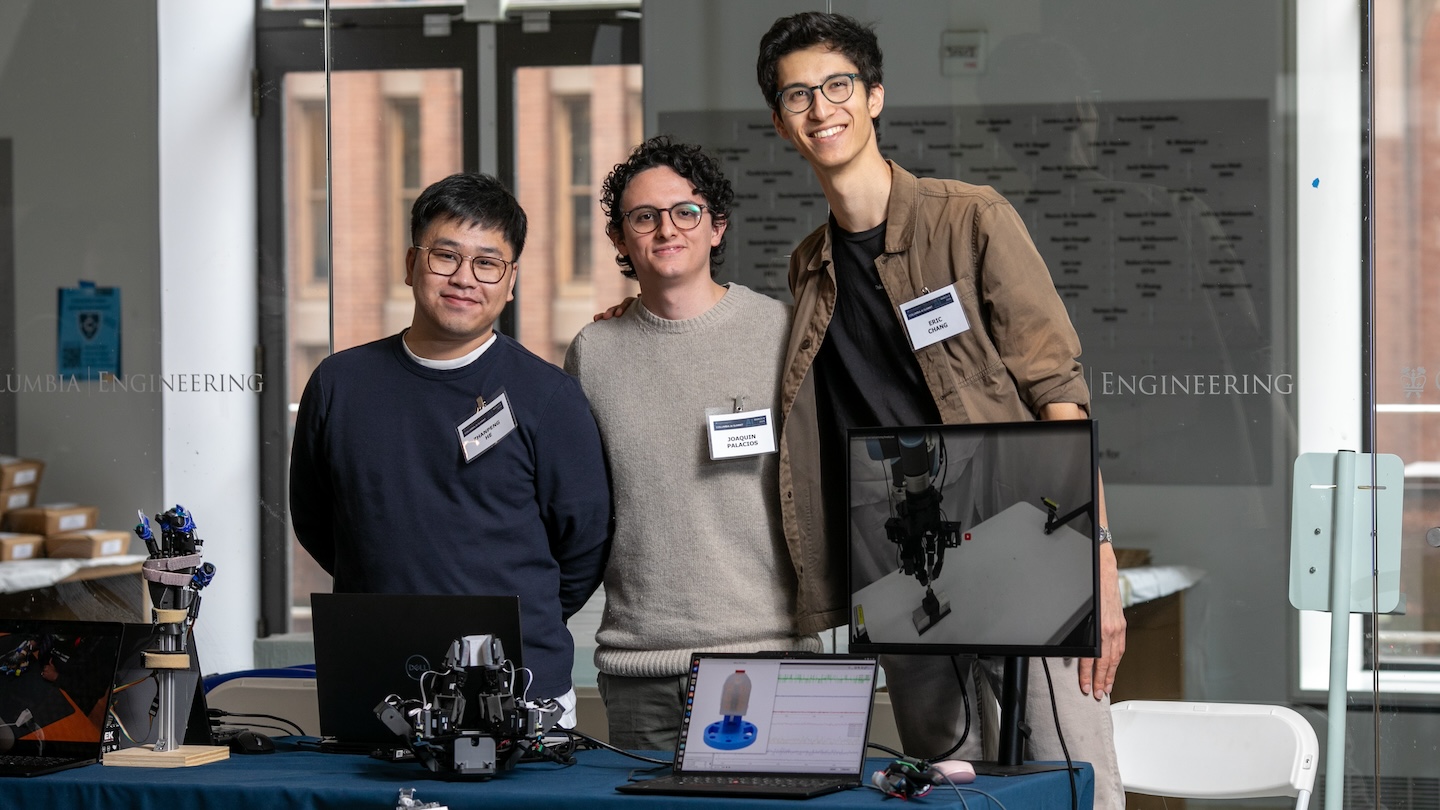 Three people standing behind a table holding a robotic hand