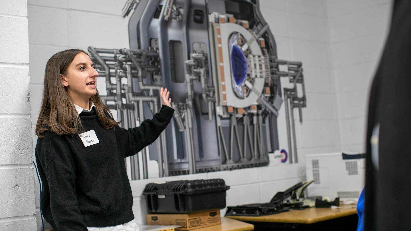 A woman gestures at a large poster of a machine