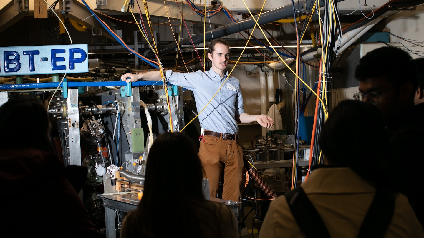 A person standing next to a large machine addressing an audience. Cables and wires are strewn in front of the speaker