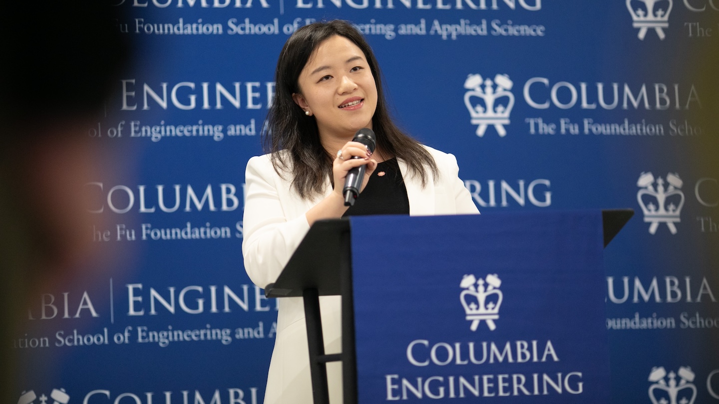 A person standing at a podium and holding a microphone. The podium and backdrop feature the Columbia Engineering logo