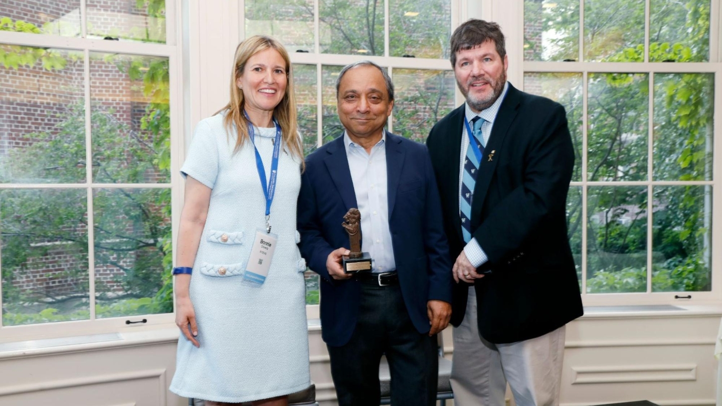 Bonnie Silvera, Hardeep Johar, and Dan Libby posing for a photo in front of a window looking out at trees. Hardeep holds the Great Teacher Award trophy