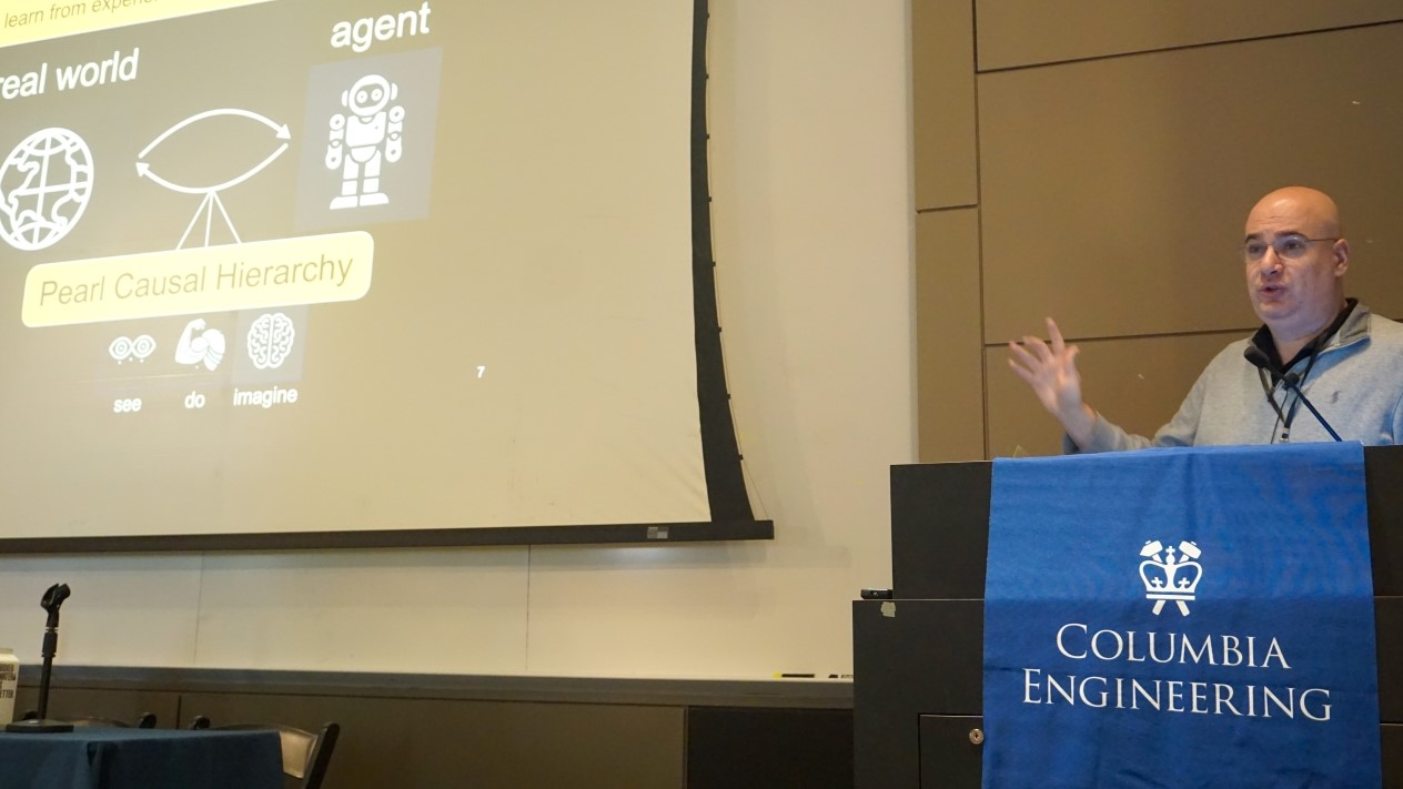 Elias Bareinboim speaking into a microphone at a podium with a Columbia Engineering banner. The screen to his right reads "Pearl Causal Hierarchy"