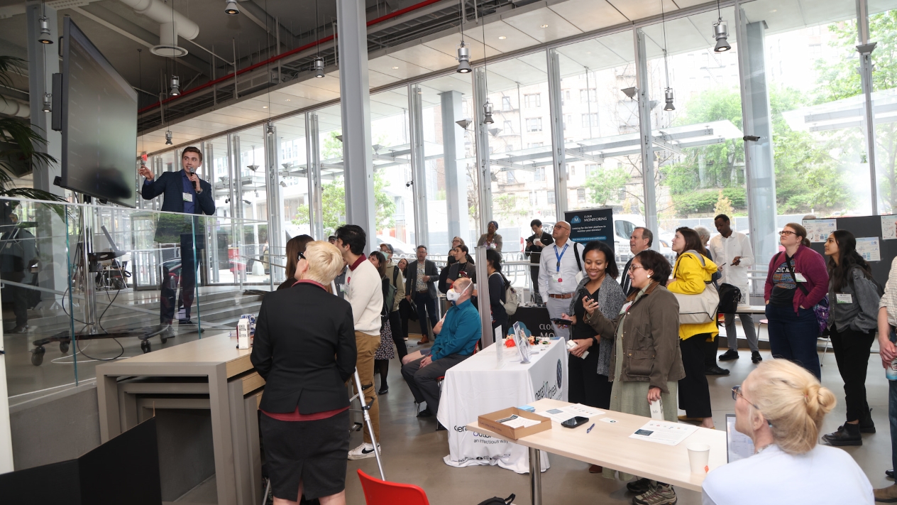 A presenter holding a microphone and pointing toward a screen while event attendees listen and mingle
