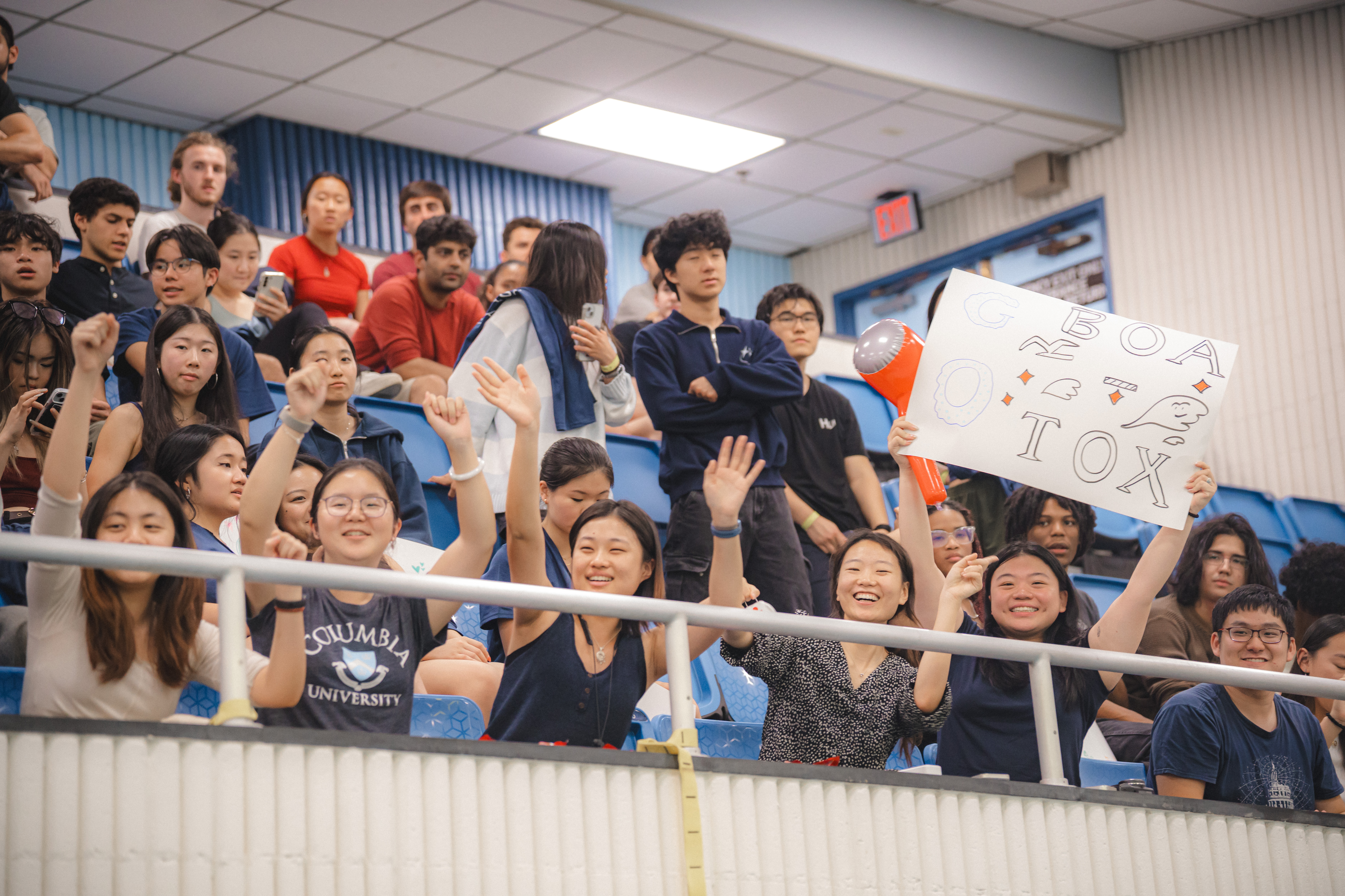 audience members in the bleachers, with one holding up a handmade "boa-tox" poster