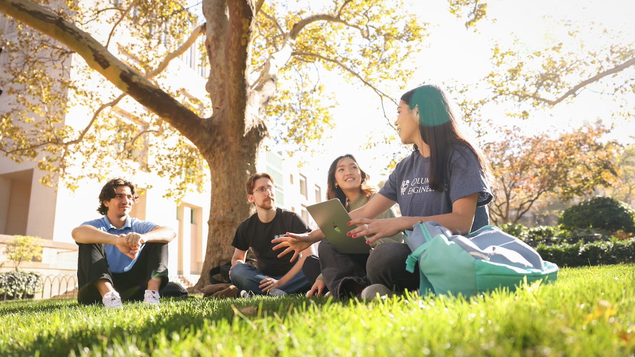 4 students seated on the grass, one with a laptop in their lap