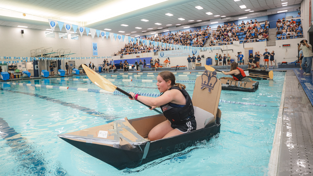 image of people rowing a small boat made of cardboard in a pool, with a large crowd watching