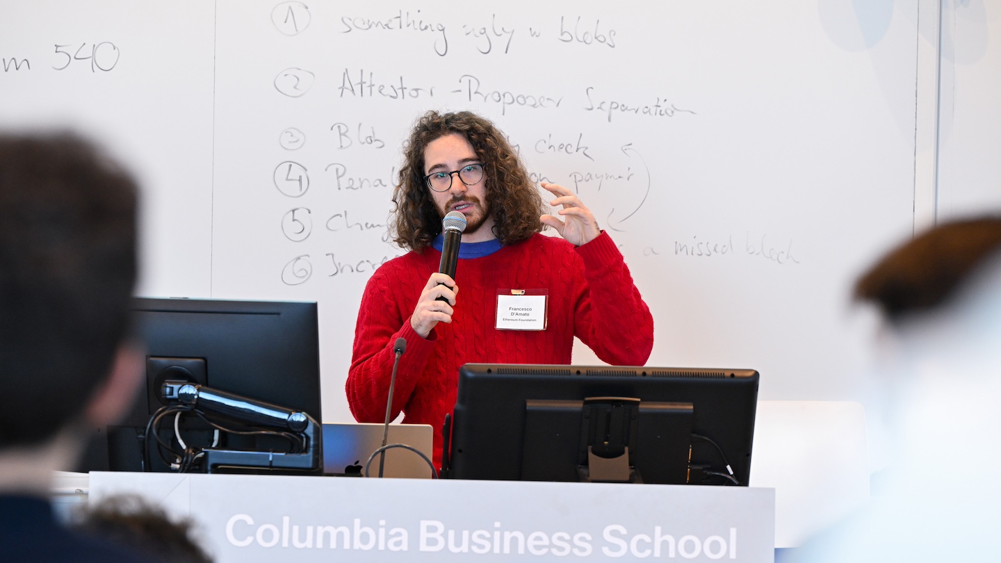 speaker at desk with microphone, with whiteboard behind