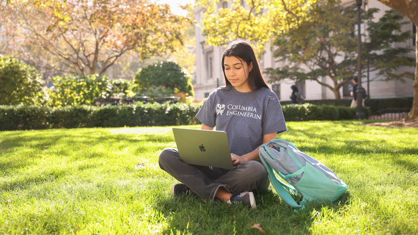 A person sitting on a sunny lawn with their laptop on their laptop. Their T-shirt reads "Columbia Engineering"