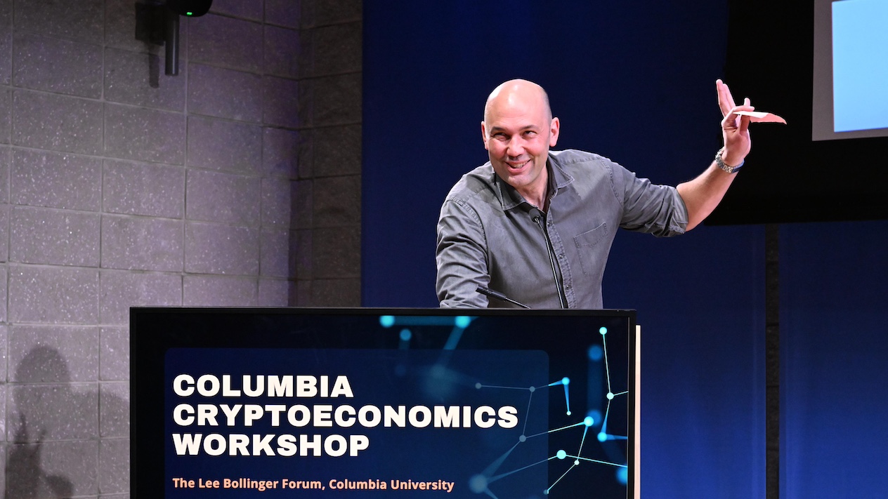 speaker at a podium, with a banner that reads "columbia cryptoeconomics workshop"
