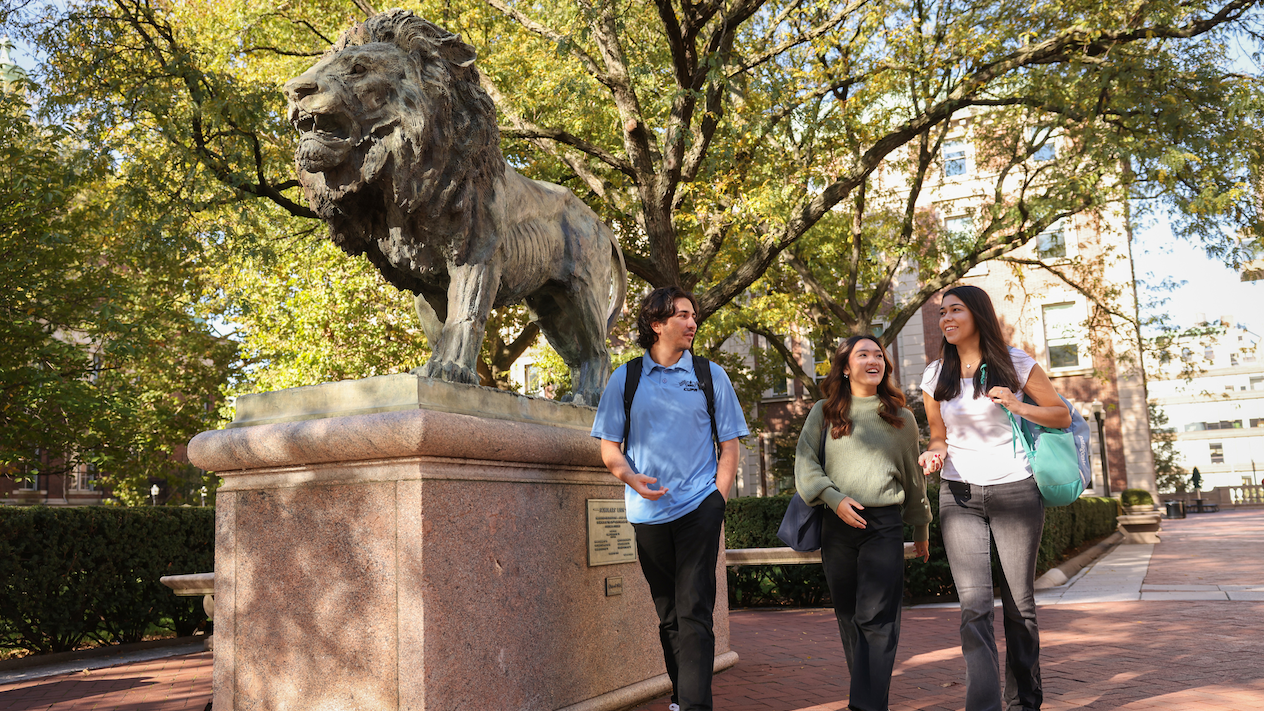 three students walking together, next to lion statue