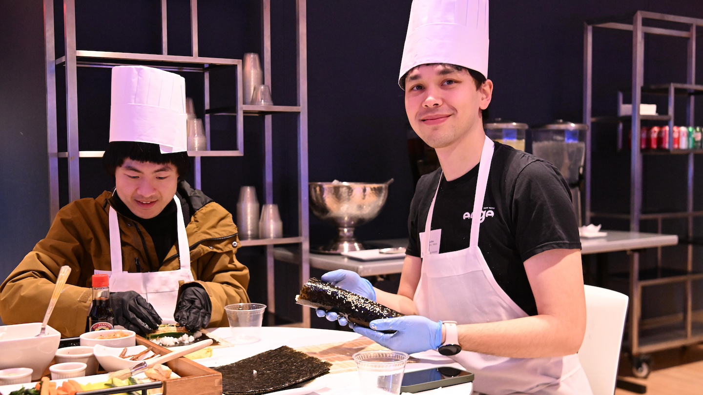 2 participants with white aprons and chefs hats making kimbap, with the participant on the right looking at the camera
