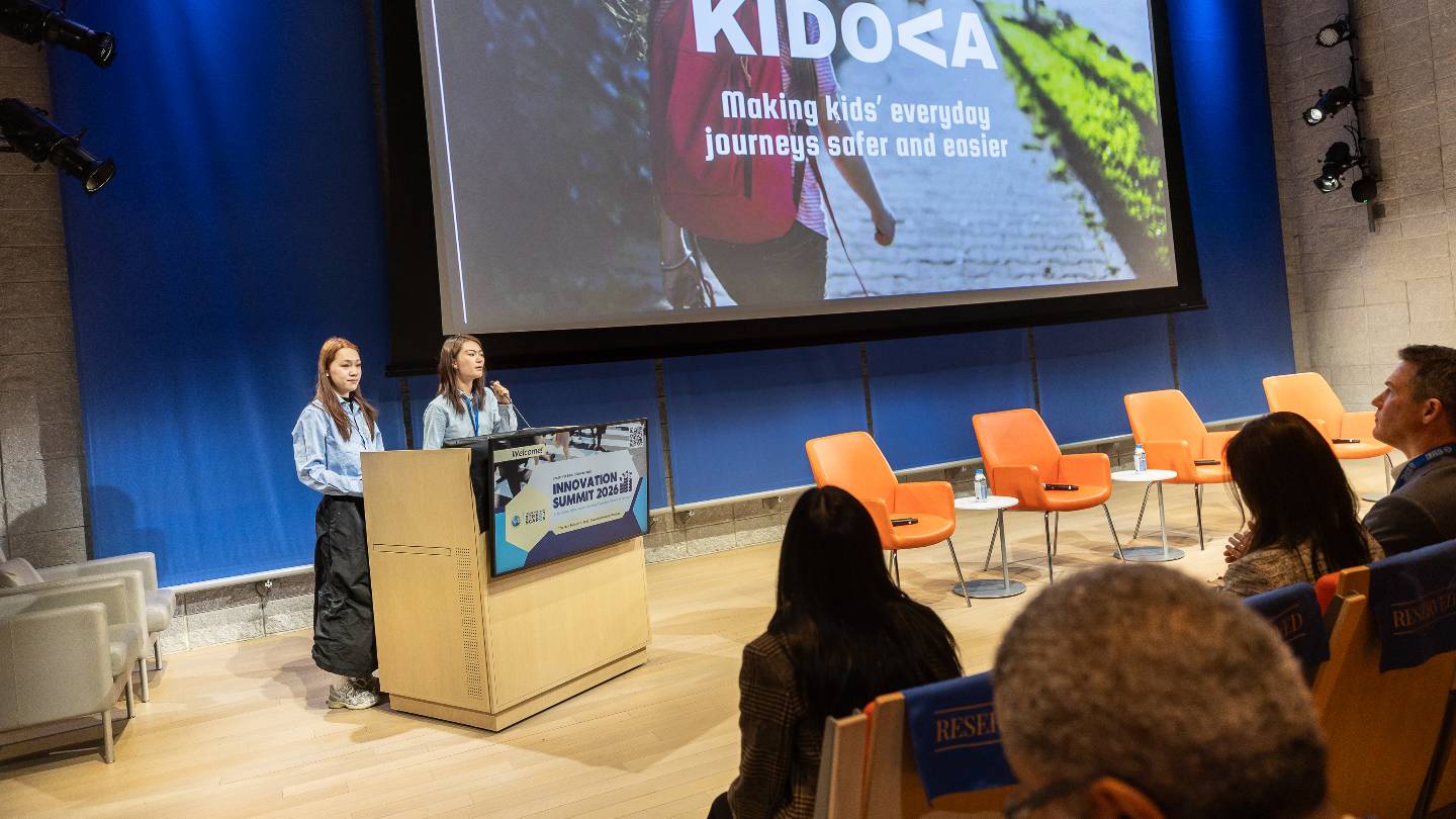 Two people standing behind a podium with a microphone on-stage. The projector screen reads "making kids' everyday journeys safer and easier"