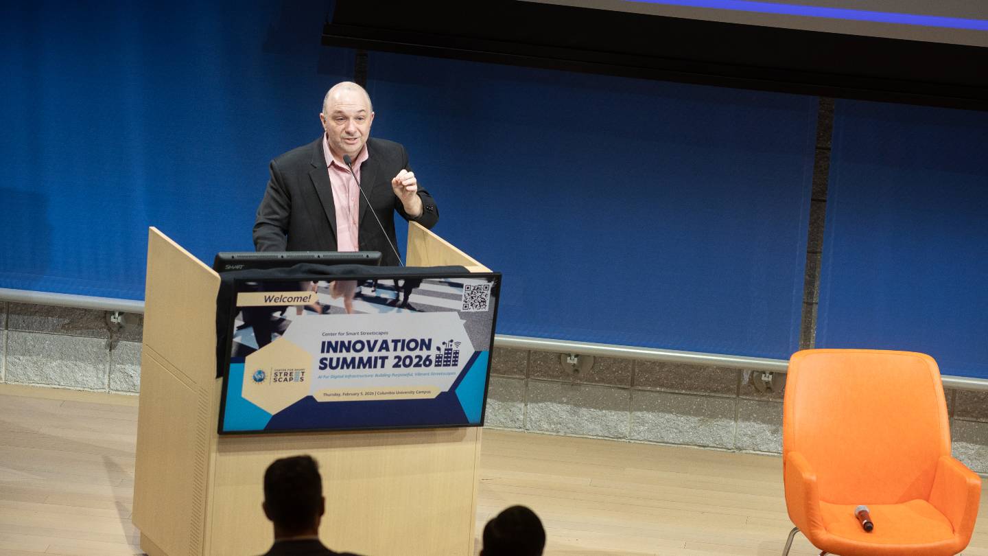 A man speaking into a microphone behind a stage podium. The sign in front of the podium reads "Welcome! Innovation Summit 2026"