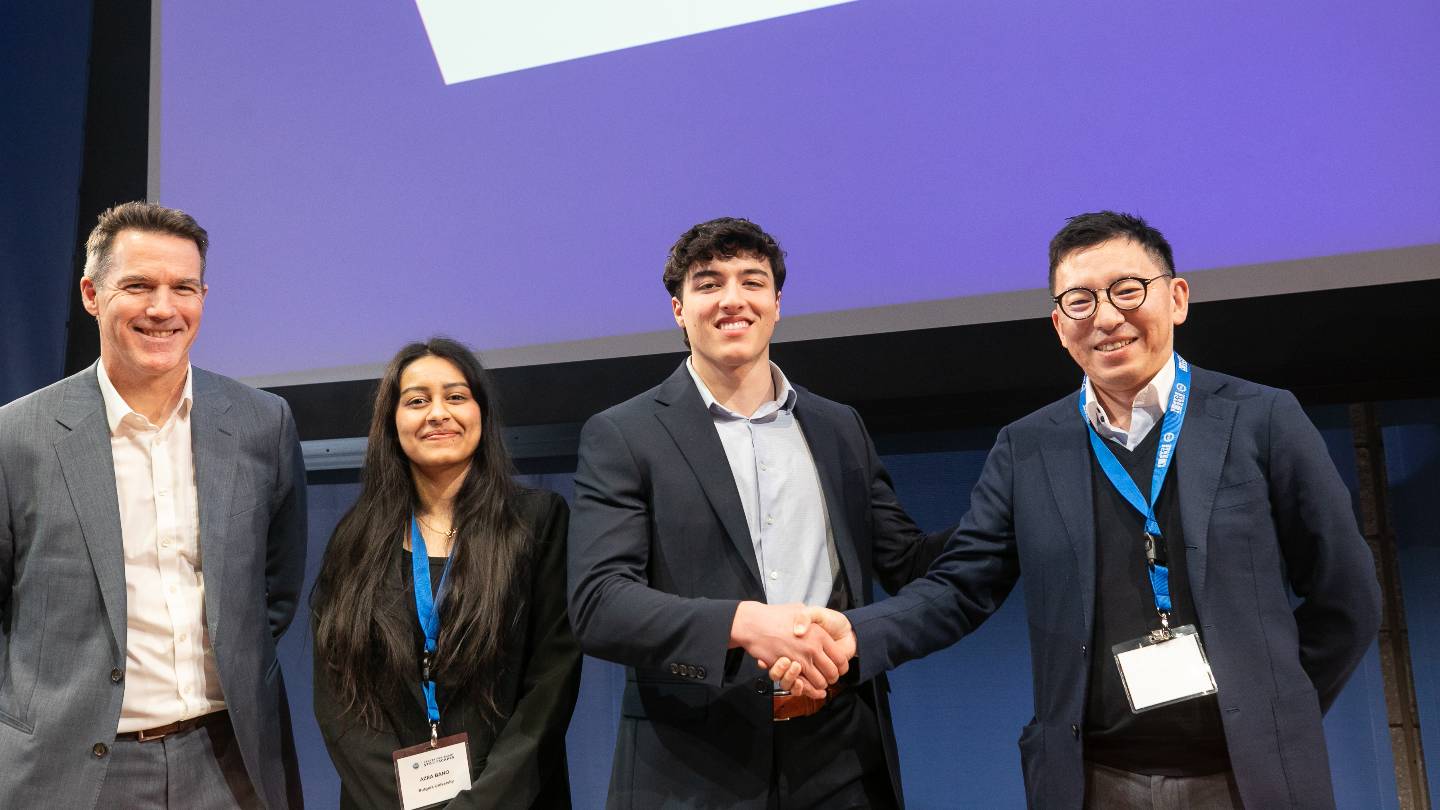 Four people posing for a photo on stage. The two people on the right shake each other's hands.