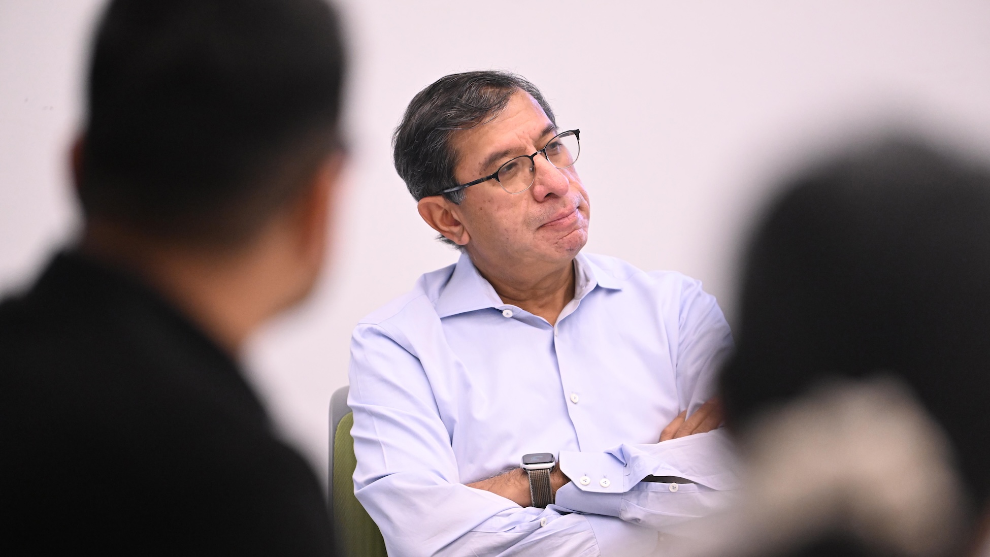 A man sits with his arms crossed, listening thoughtfully in a meeting; two blurred people frame the foreground.