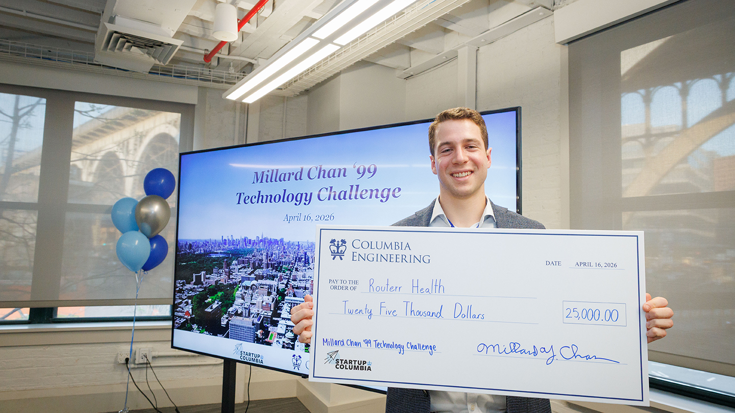 Smiling competition winner holds a large $25,000 Columbia Engineering check in front of a screen reading “Millard Chan ’99 Technology Challenge,” with blue and silver balloons nearby.