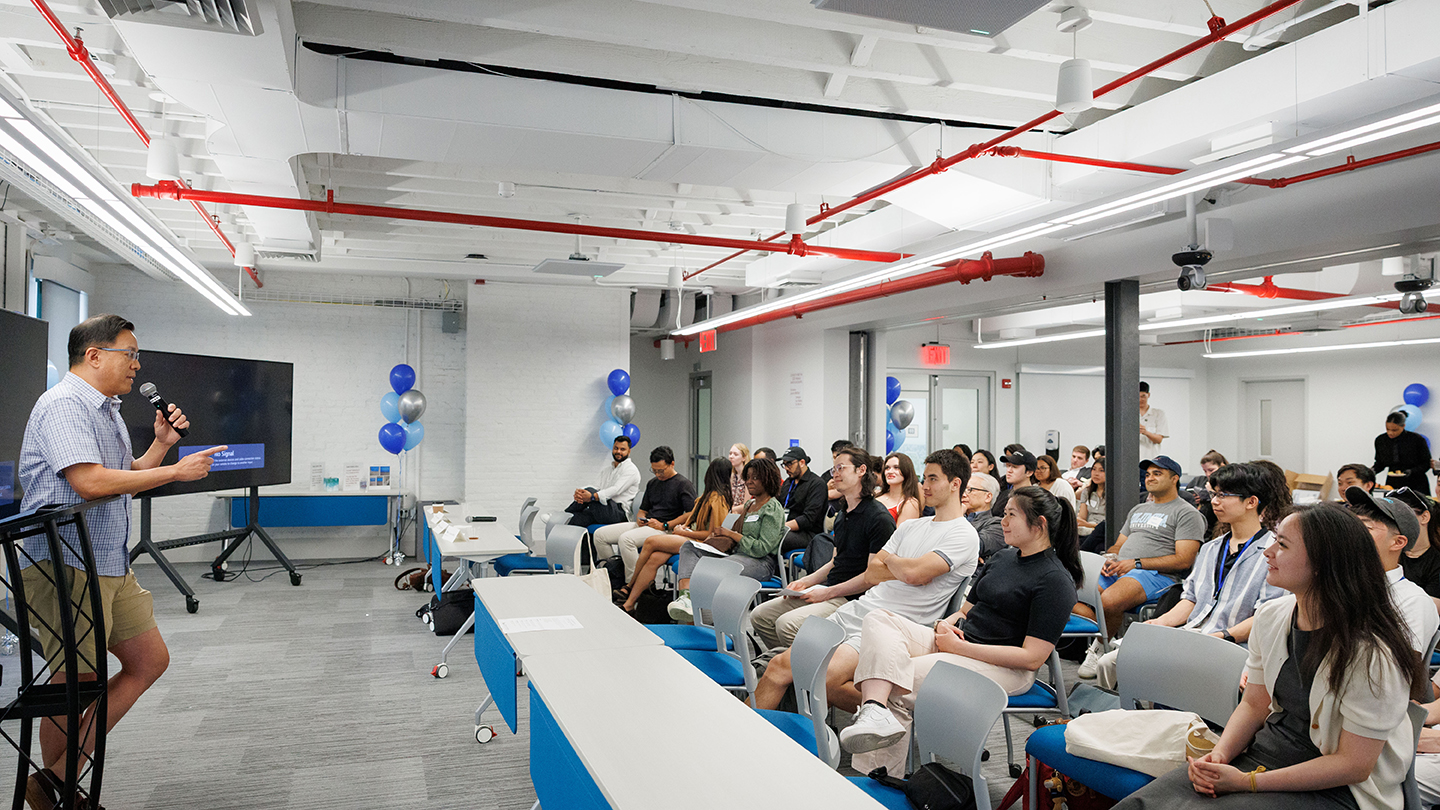 Speaker at a podium addresses an audience seated in a bright event room decorated with blue and silver balloons.