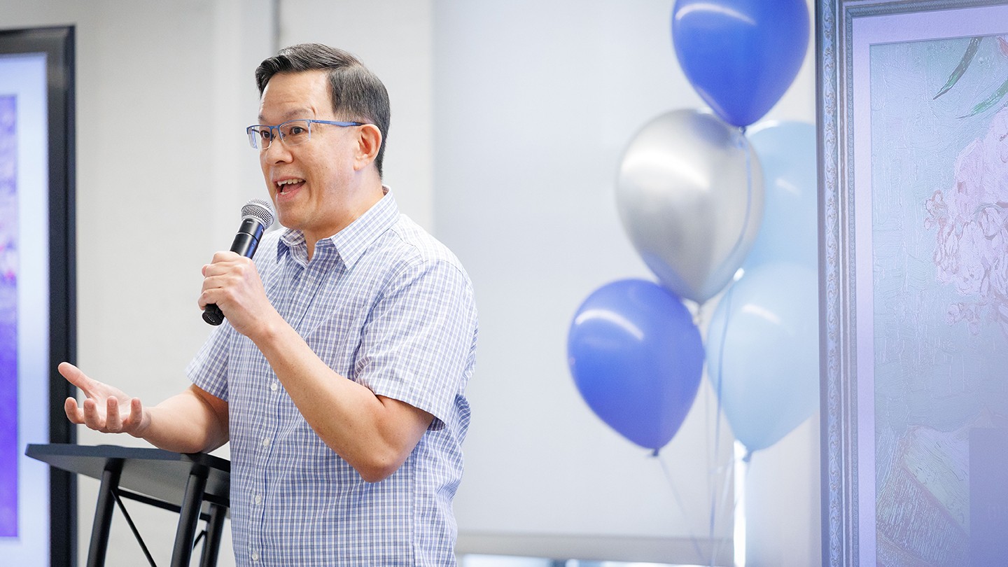 A man speaking into a handheld microphone, gesturing, with balloons and a display in the background.