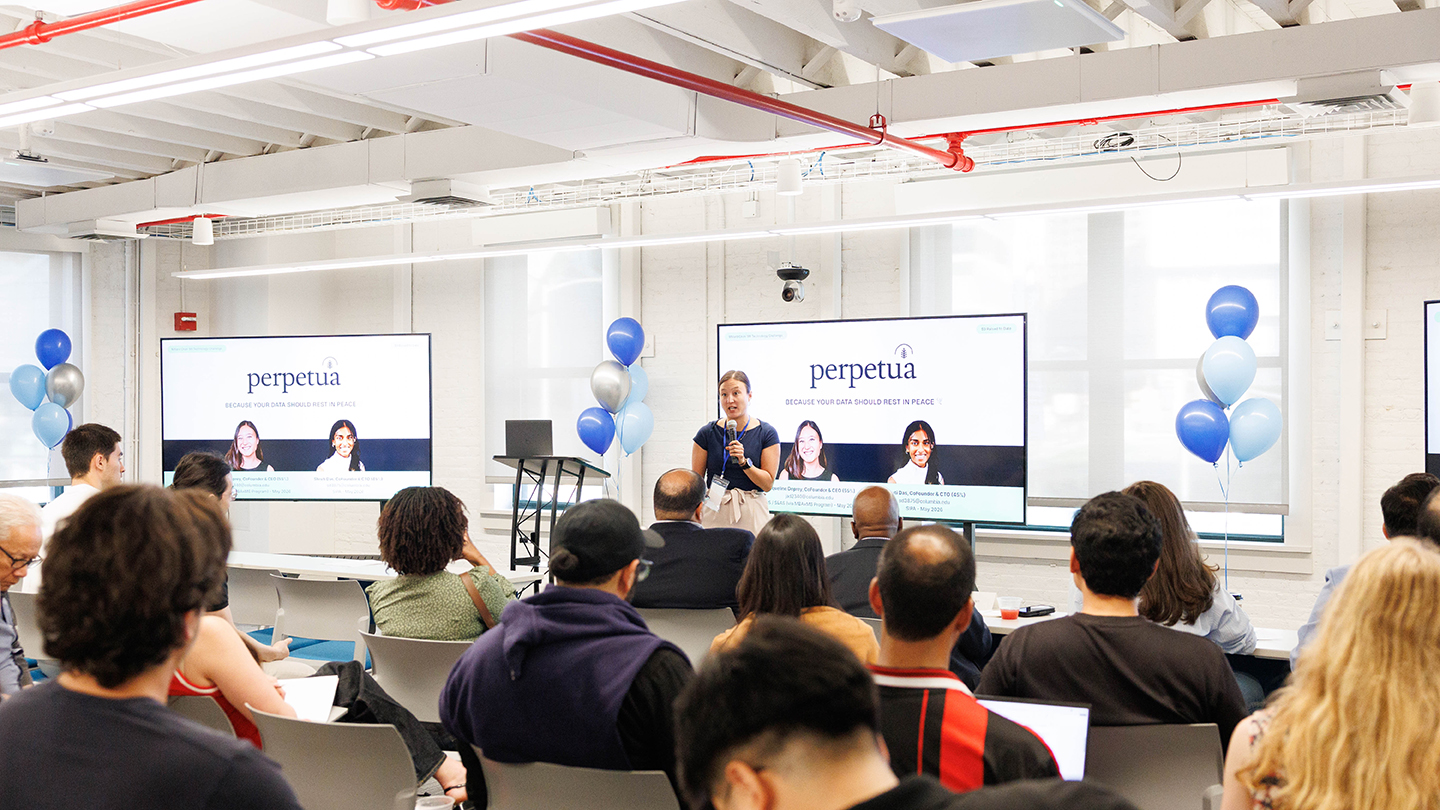 Wide shot of a presenter speaking to a seated audience; two large screens show a “perpetua” presentation slide, with balloon clusters along the walls.