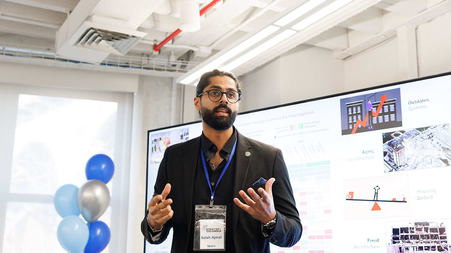 Presenter in a dark blazer gestures while speaking in front of a slide deck; blue and silver balloons stand to the side.
