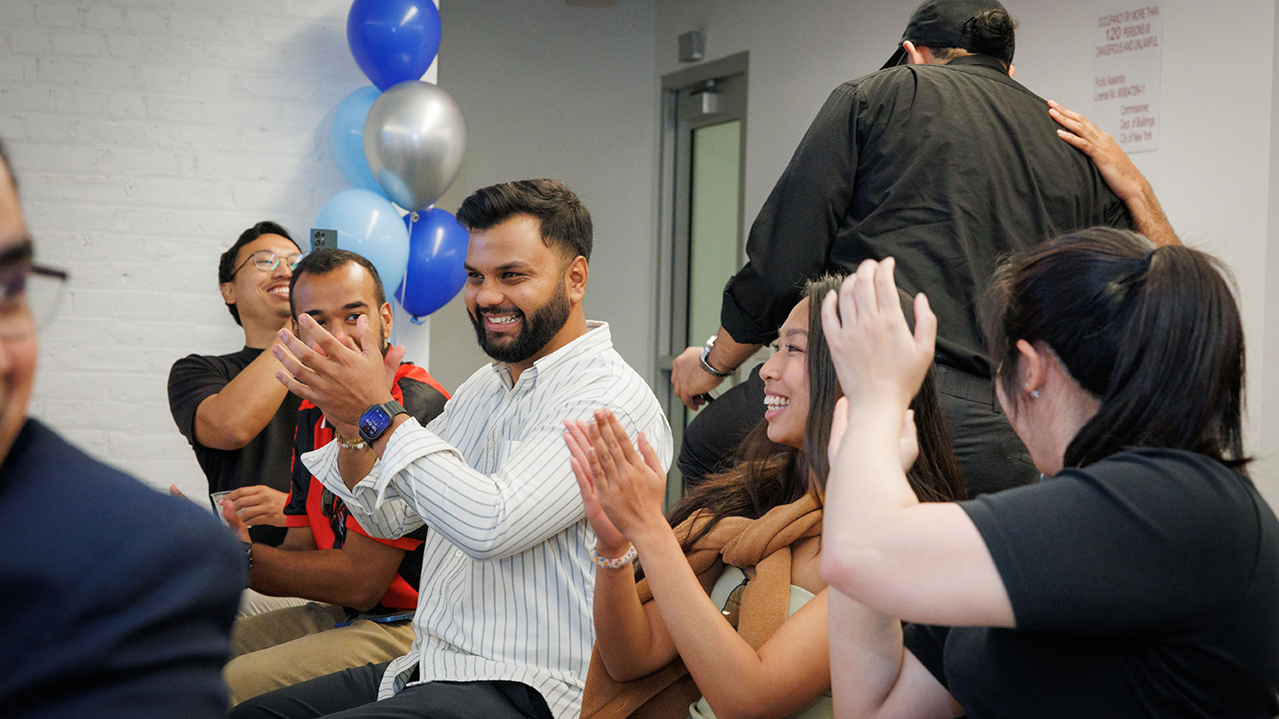 Audience members clap and smile during the event; blue and silver balloons are visible in the background.