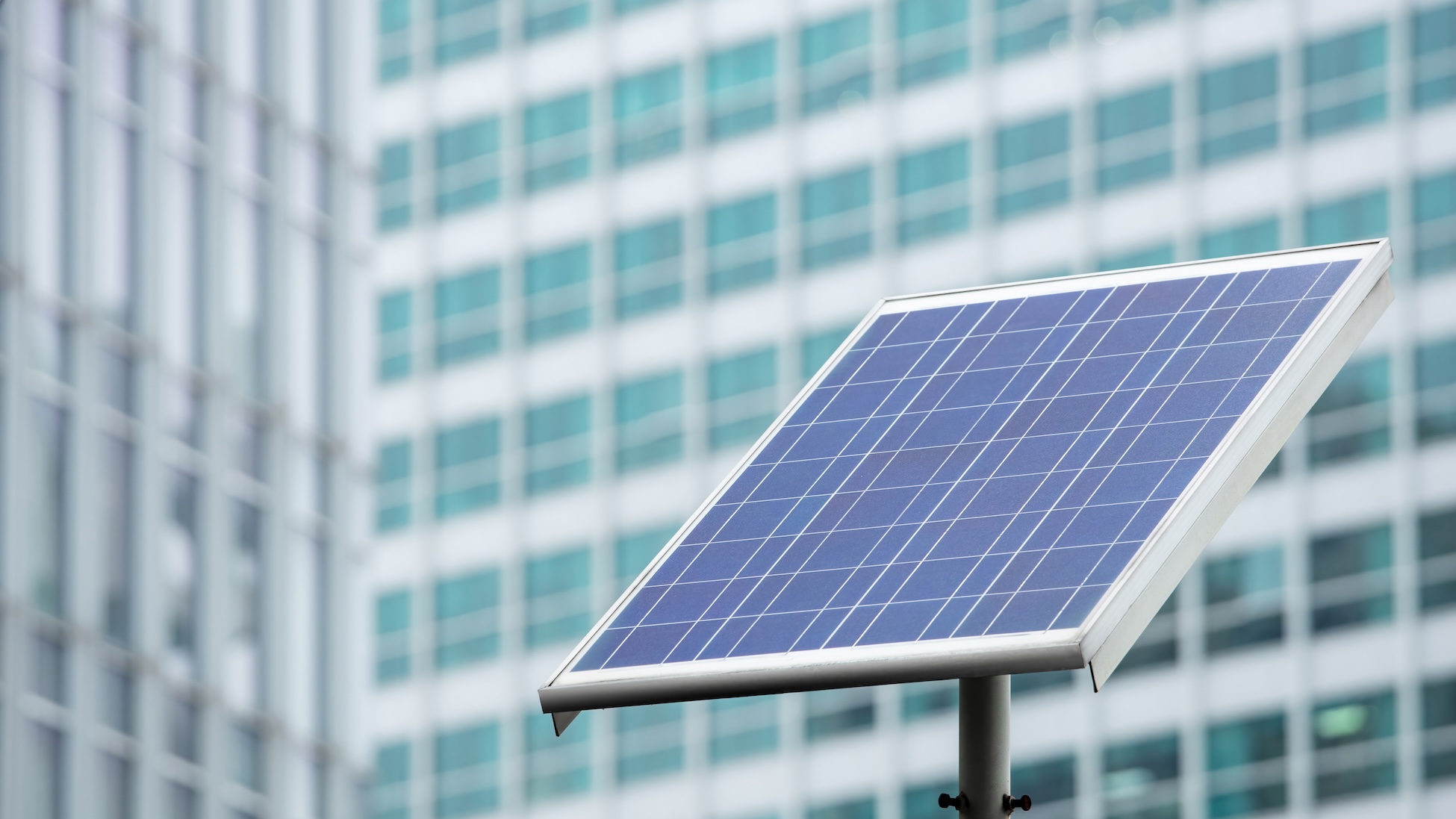 Close-up of a tilted solar panel mounted on a pole, with a blurred high-rise building of glass windows in the background.