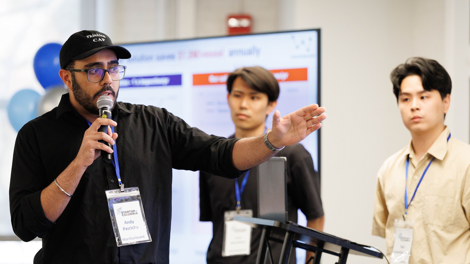  man wearing a black cap and glasses speaks into a handheld microphone and gestures with his arm extended during a presentation; two other participants with name badges stand nearby, with a large screen and blue-and-white balloons in the background.