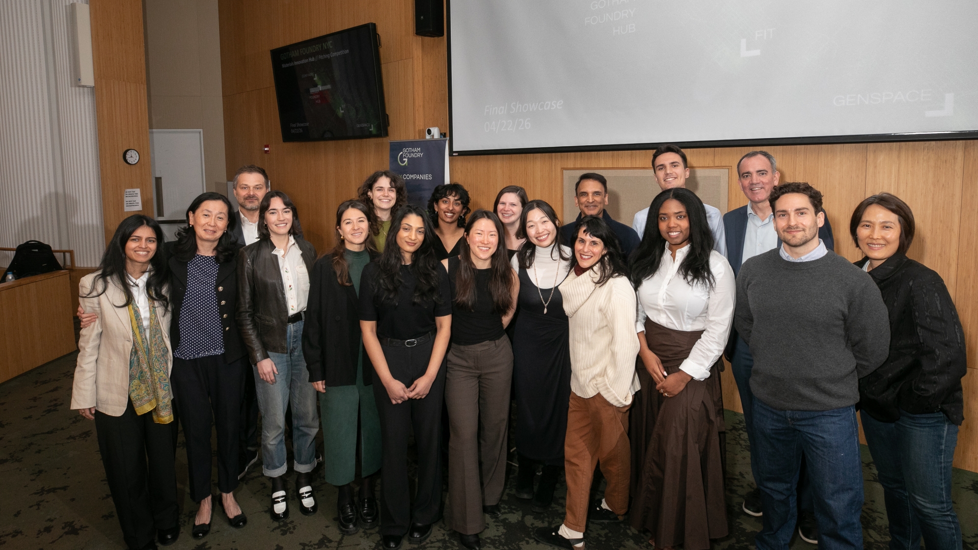 A group of about 16 adults pose together in an event space, smiling at the camera. They stand in two rows in front of a large projector screen and a small banner (with “Gotham Foundry” visible) on a wood-paneled wall.